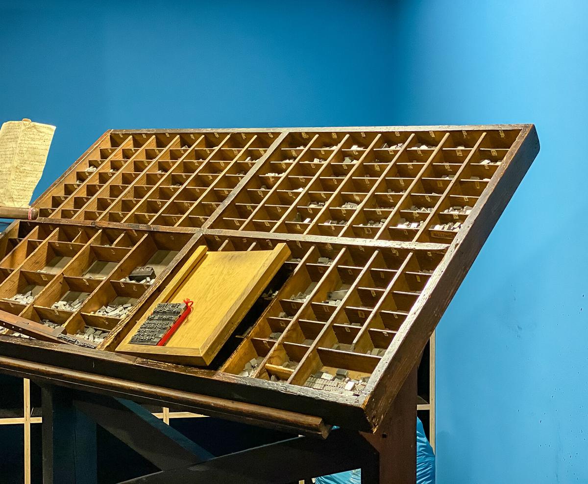 Casse typographique en bois inclinée, garnie de caractères d’imprimerie en plomb rangés par compartiments, avec un composteur posé au centre.